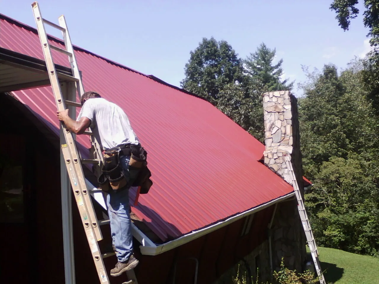 Roofer on a ladder beside a red metal roof and stone chimney — Soffit & Fascia Repair by Roofing Brothers in Arendtsville